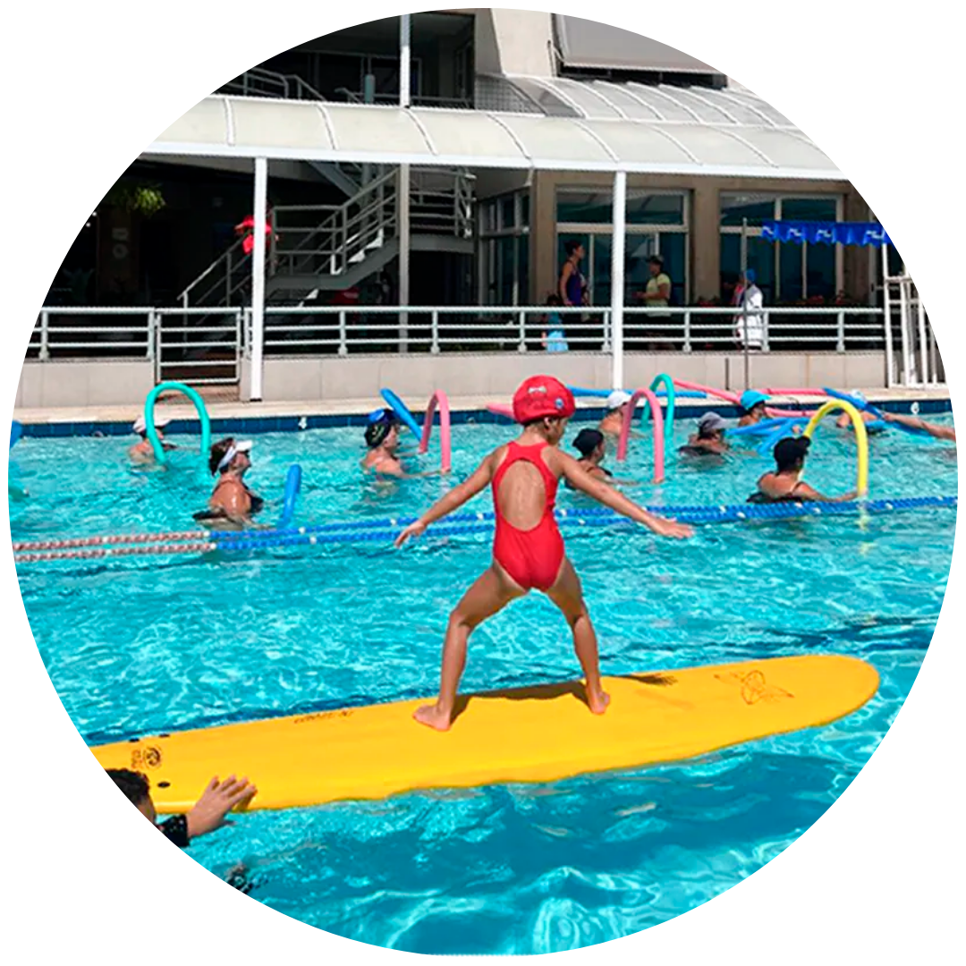 Criança a equilibrar-se numa prancha de surf grande na piscina durante uma atividade lúdica complementar da natação infantil na academia Acqua Fitness, no Recreio dos Bandeirantes.